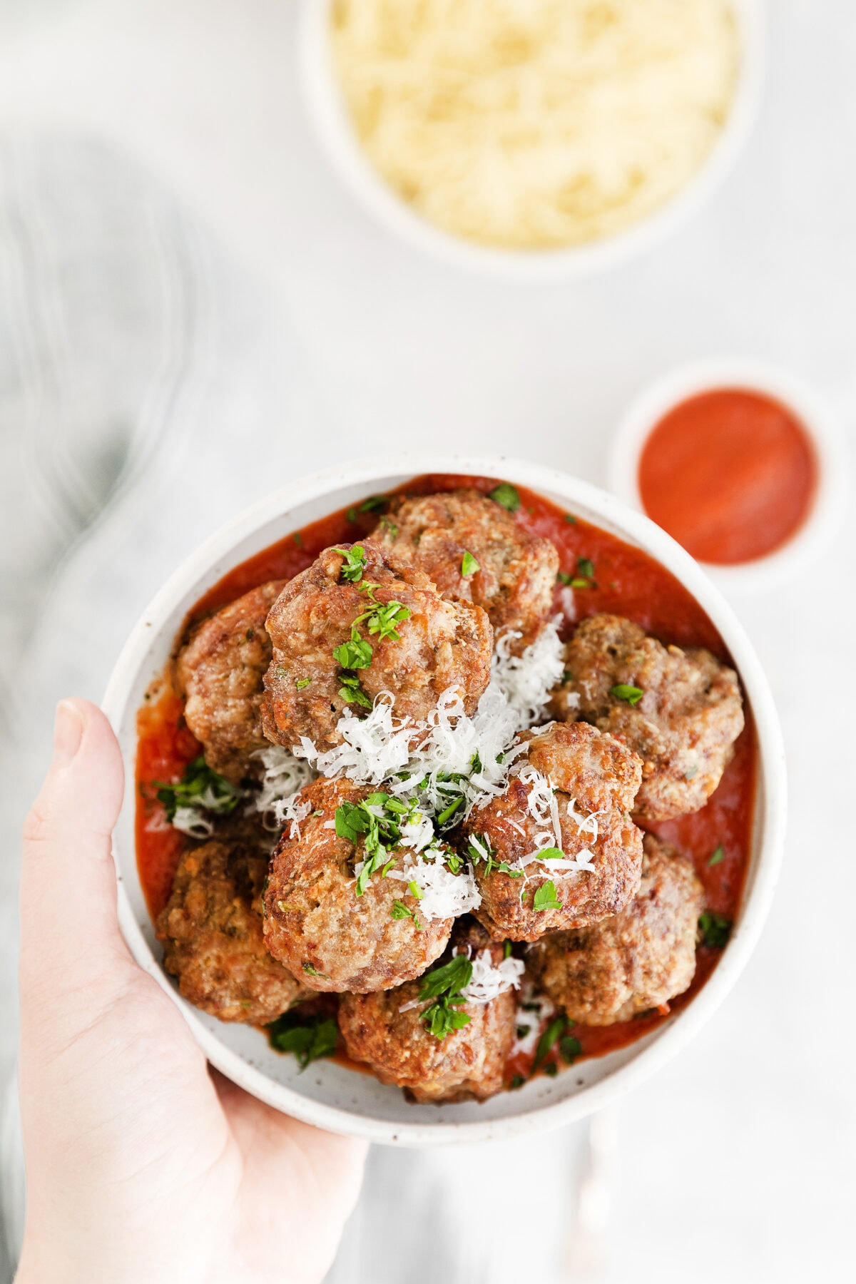 air fryer meatballs in a white bowl being held by a hand with a bowl of sauce and pasta in the background.