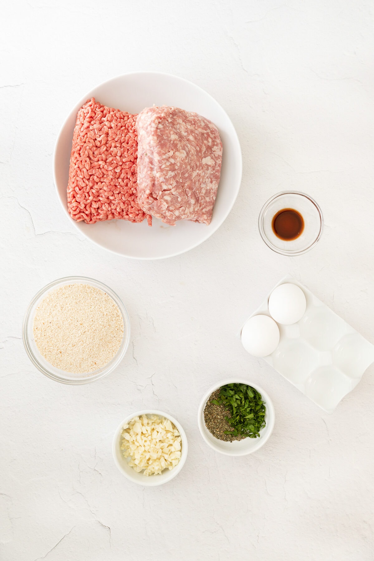 raw meat on a plate surrounded by bowls for of ingredients for meatballs like herbs, cheese, breadcrumbs, eggs.