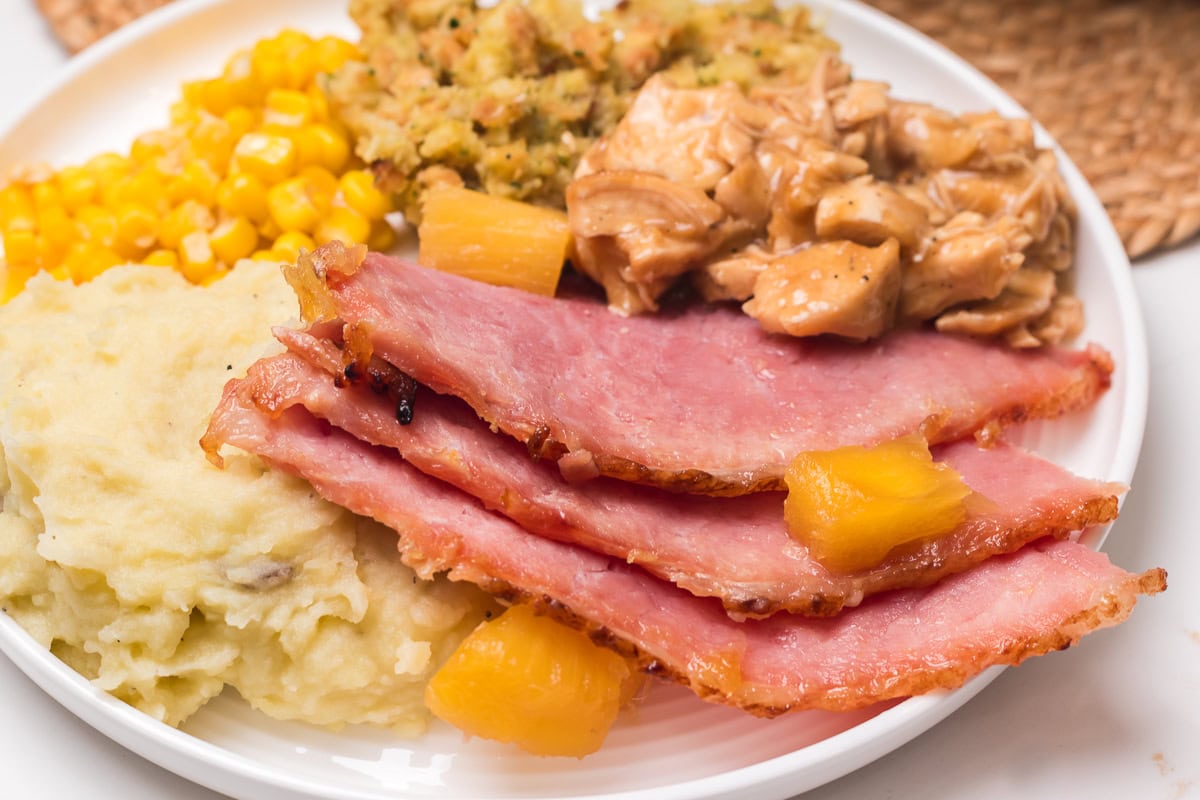 close up picture of plate of thanksgiving dinner with baked ham, turkey and gravy, buttered corn, mashed potatoes and stuffing.