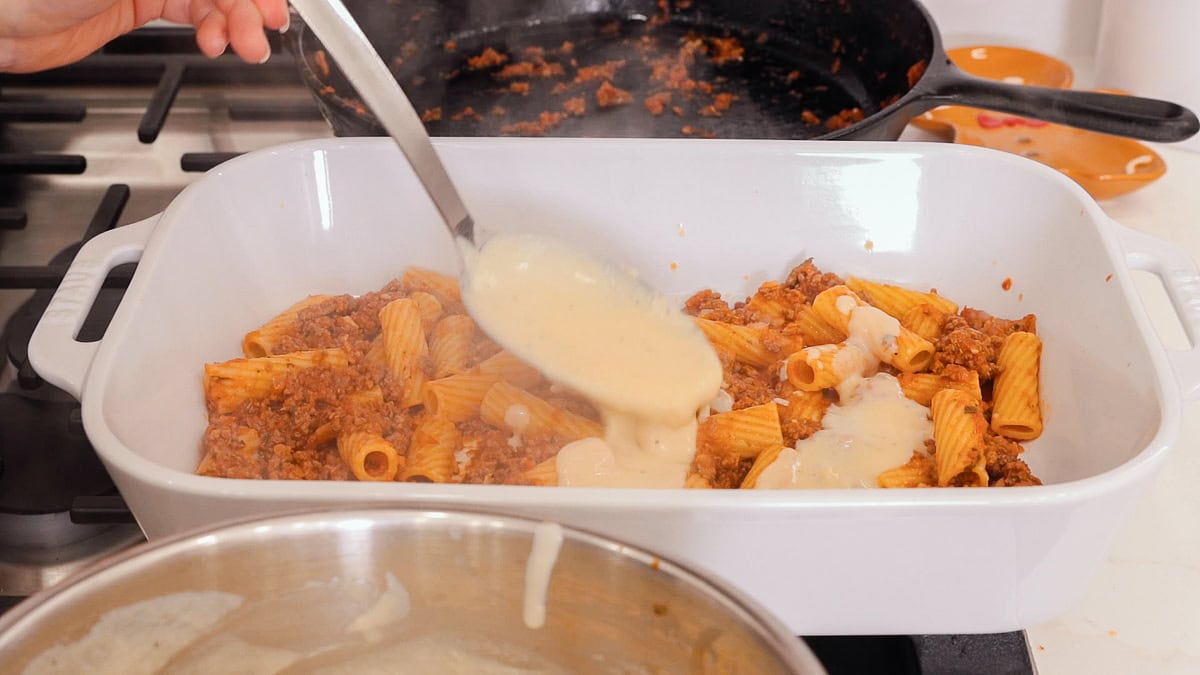 meat sauce with rigatoni in casserole dish with bechamel being ladled over with a spoon.