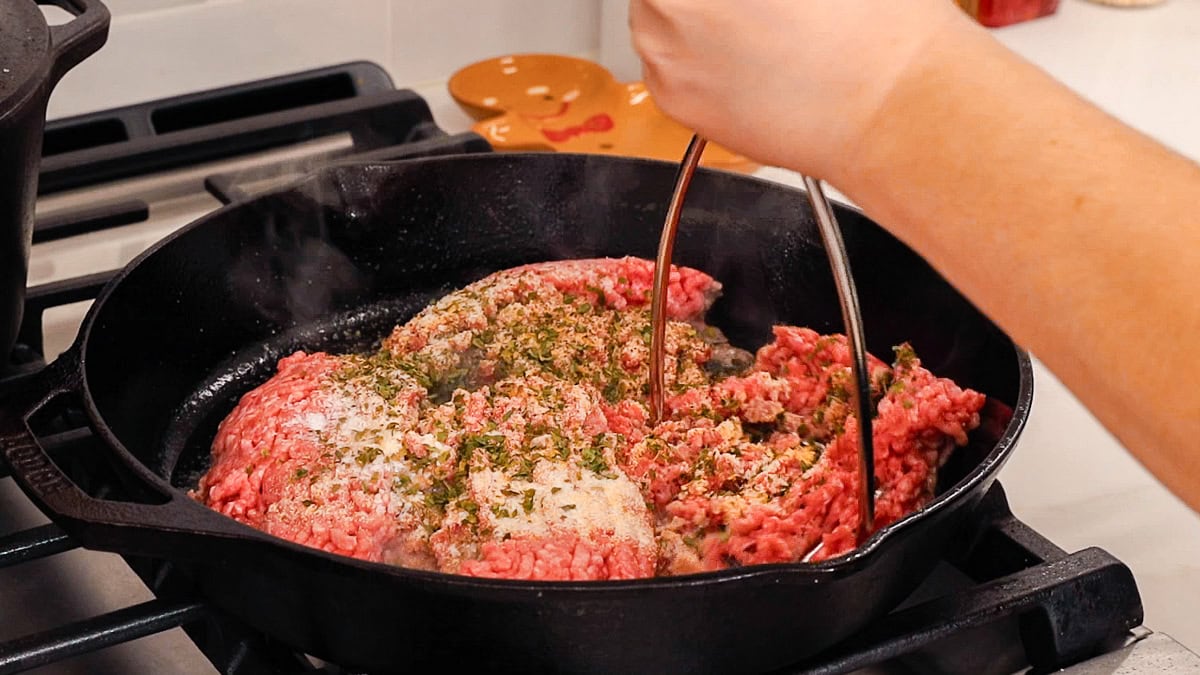 seasoned ground beef cooking in cast iron skillet with a hand holding a potato masher to mash and cook the beef.