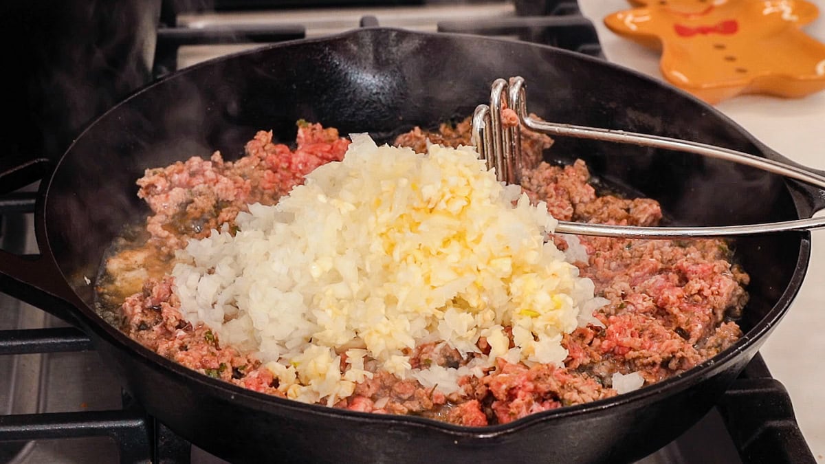 partially cooked ground beef in a skillet with raw onion and raw garlic ready to be cooked on top.