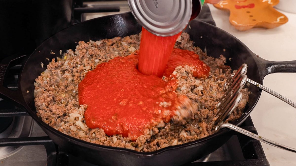 adding a can of crushed tomatoes to a skillet with ground beef in it.