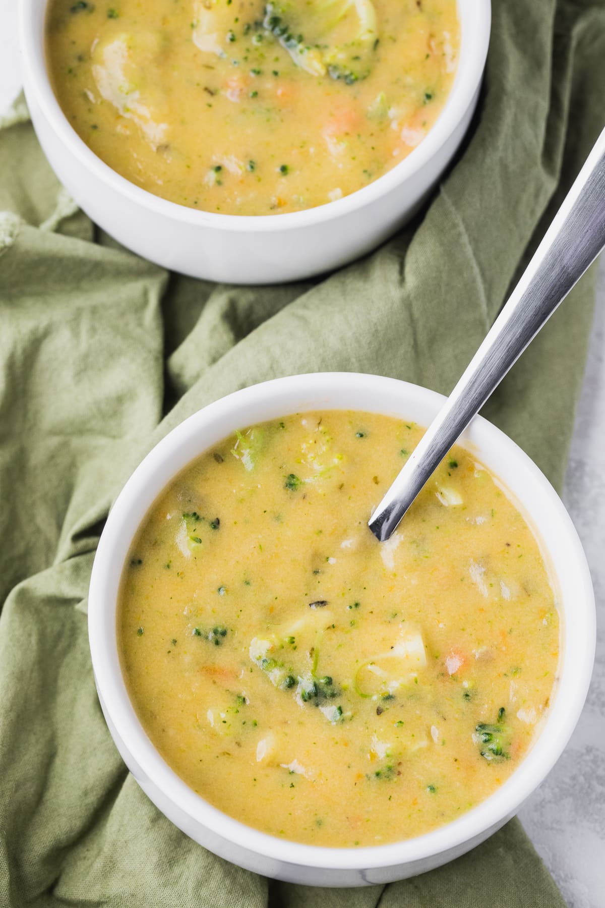 overhead photo of broccoli potato soup in 2 white bowls on a green napkin with spoon in bowl.