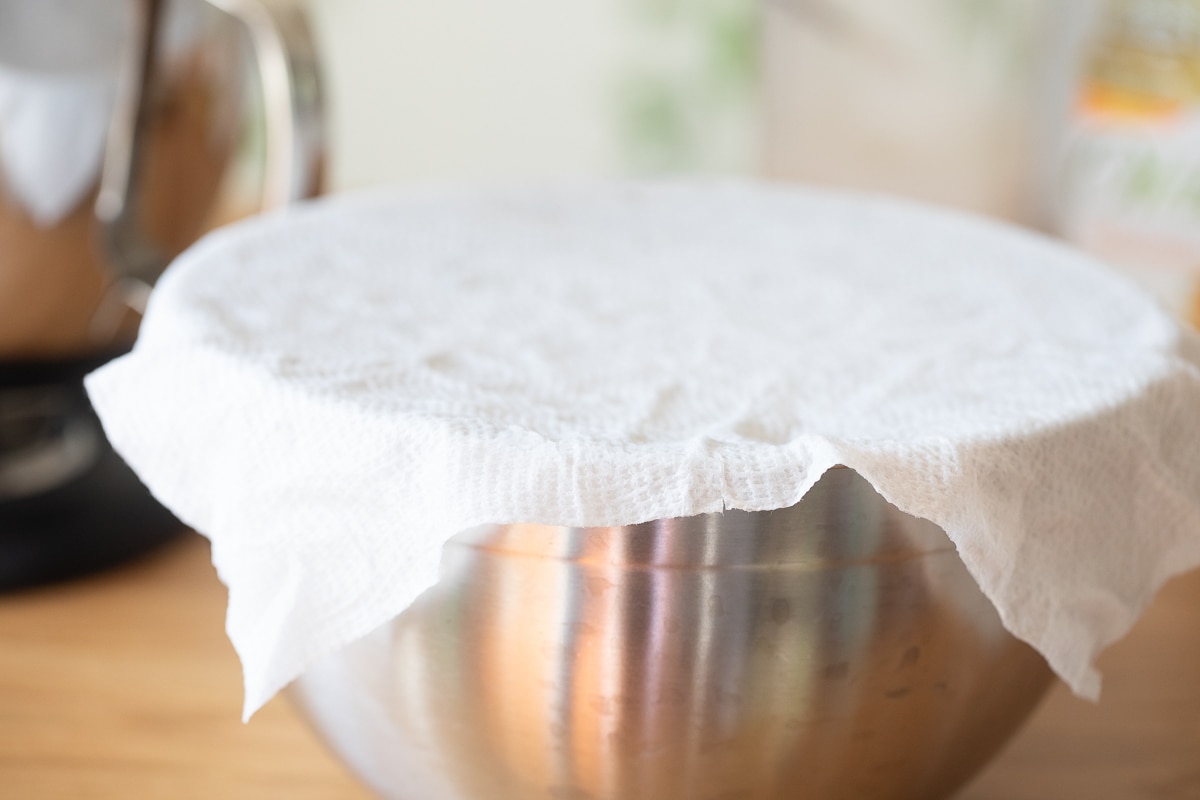 donut dough in a stainless steel bowl with a piece of paper towel over top of it.