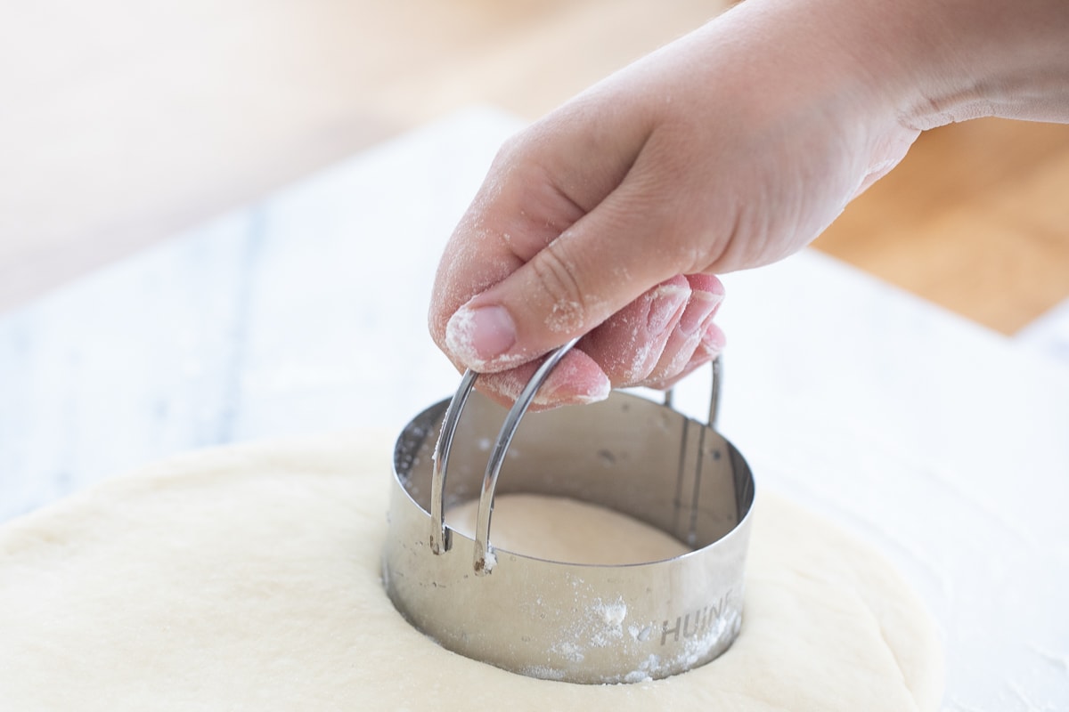 hand using a biscuit cutter to make donuts on a marble slab.