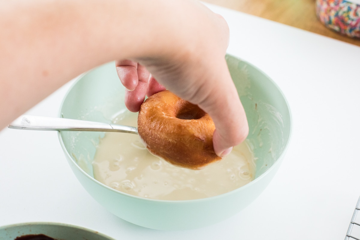 hand dipping donut into glaze in a light green bowl with a spoon in it.