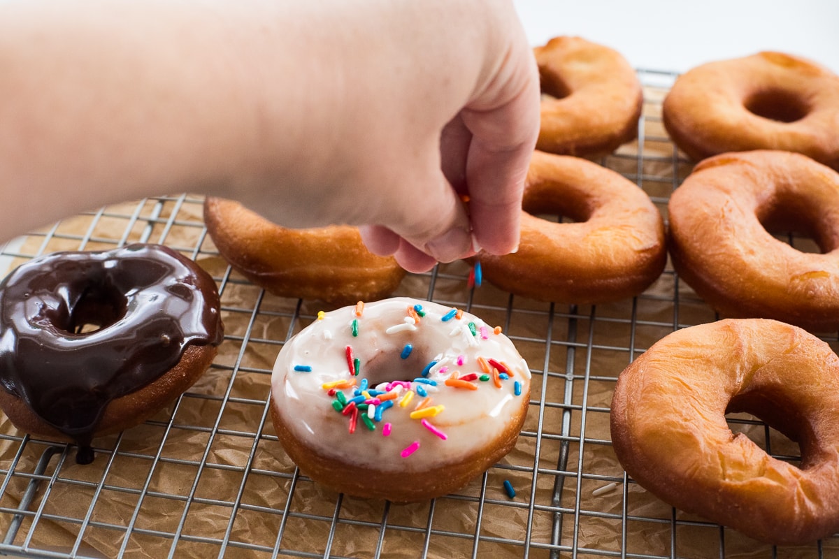 hand sprinkling sprinkles onto donut on a rack.