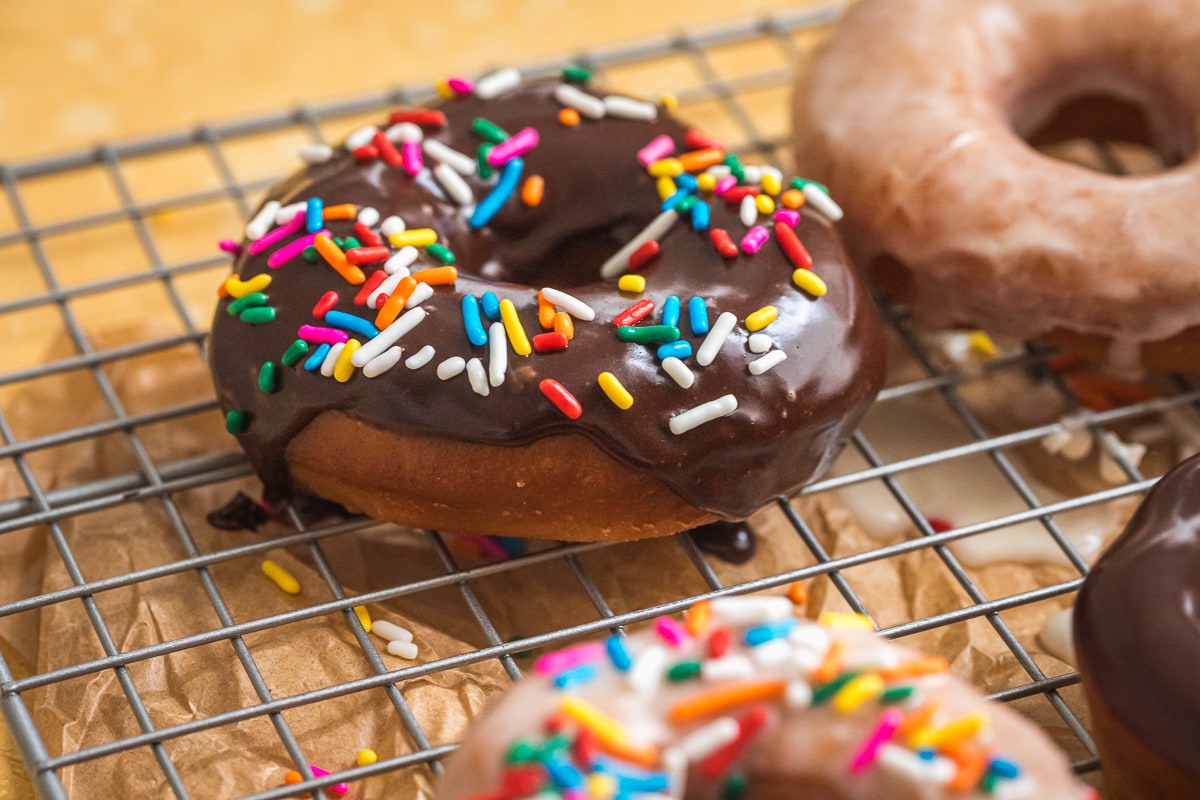 close up of chocolate glazed donut with rainbow sprinkles on a cooling rack.