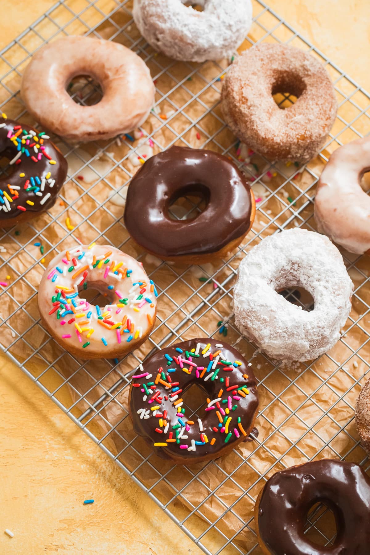 donuts with sprinkles, glaze, powdered sugar, and cinnamon sugar on a cooling rack on a yellow backdrop.