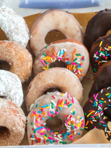 dairy free donuts in a bakery box covered in chocolate glaze, some in vanilla glaze, some with cinnamon sugar, and some with powdered sugar.