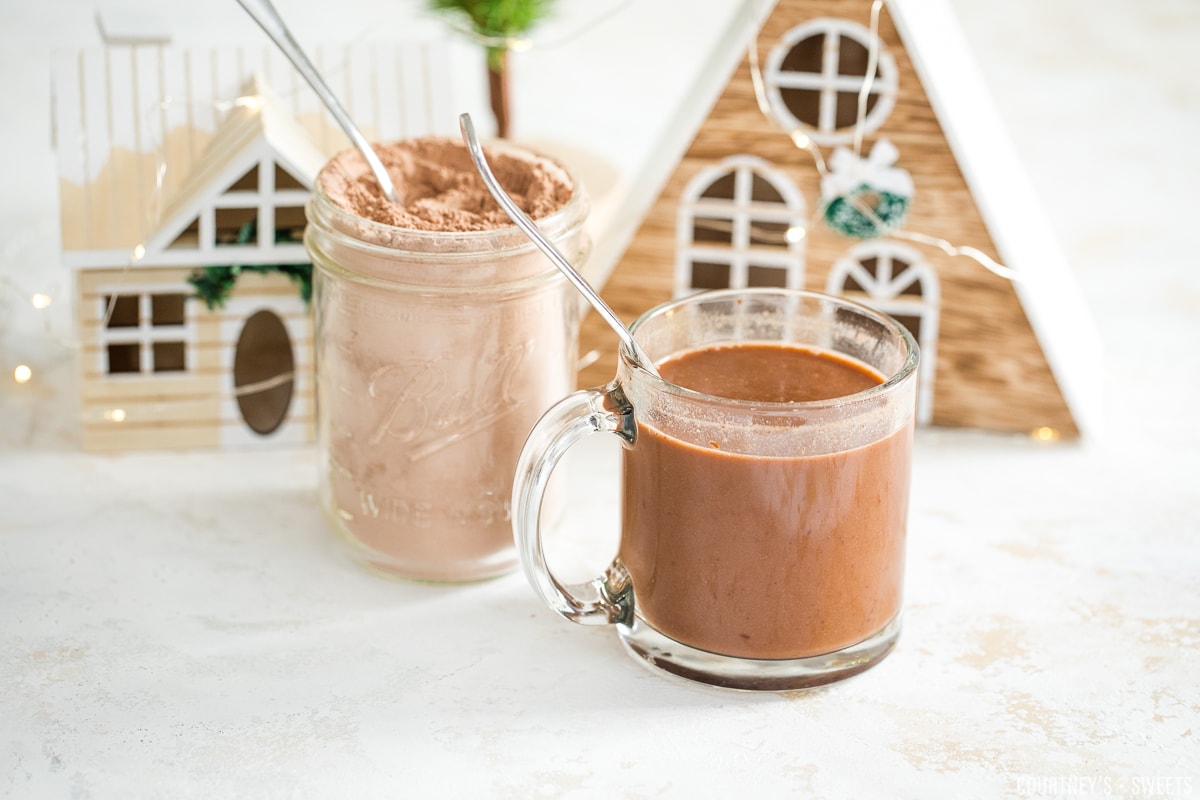 dairy free hot chocolate mix in a mason jar and mixed into plant milk in a glass mug with little christmas houses behind them