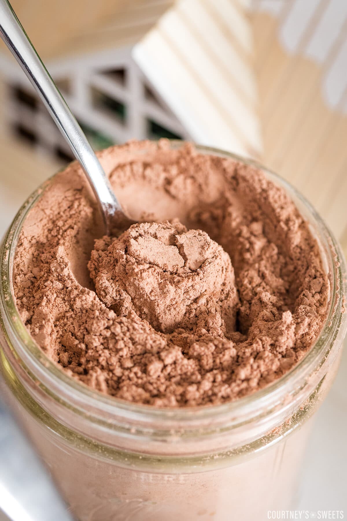 close up of dairy free hot chocolate mix in a mason jar with spoon inside of it with little wooden house top in background