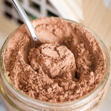 close up of dairy free hot chocolate mix in a mason jar with spoon inside of it with little wooden house top in background