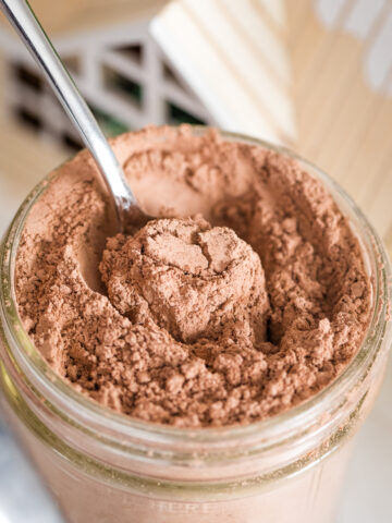 close up of dairy free hot chocolate mix in a mason jar with spoon inside of it with little wooden house top in background