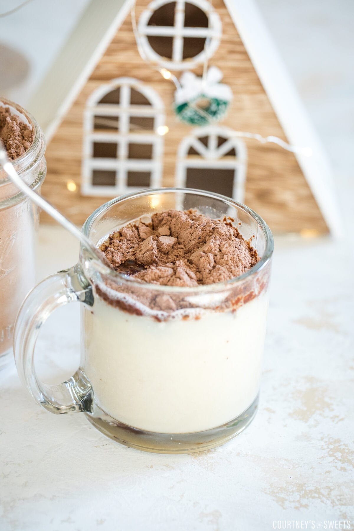 dairy free hot chocolate mix in a mason jar and scooped into plant milk in a glass mug with little christmas houses behind them