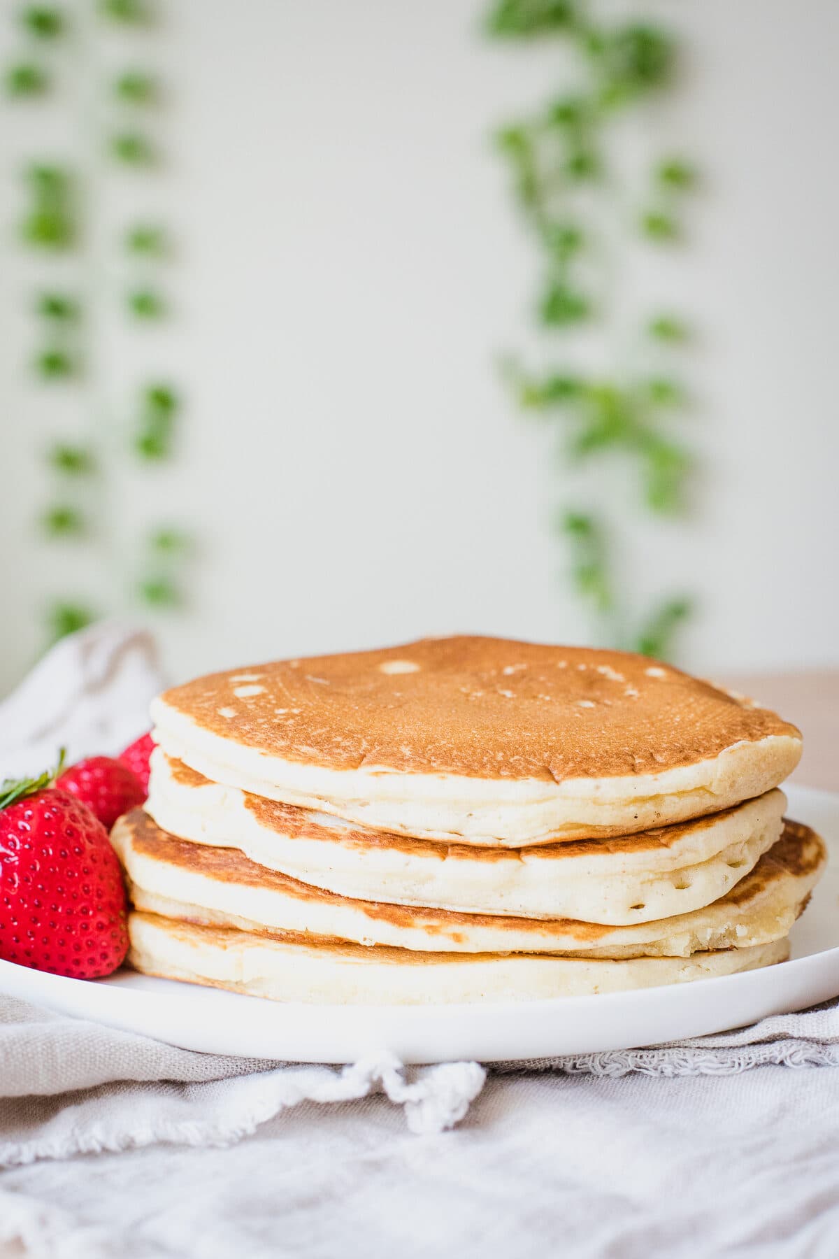 stack of dairy free pancakes on a white plate with greenery in background.