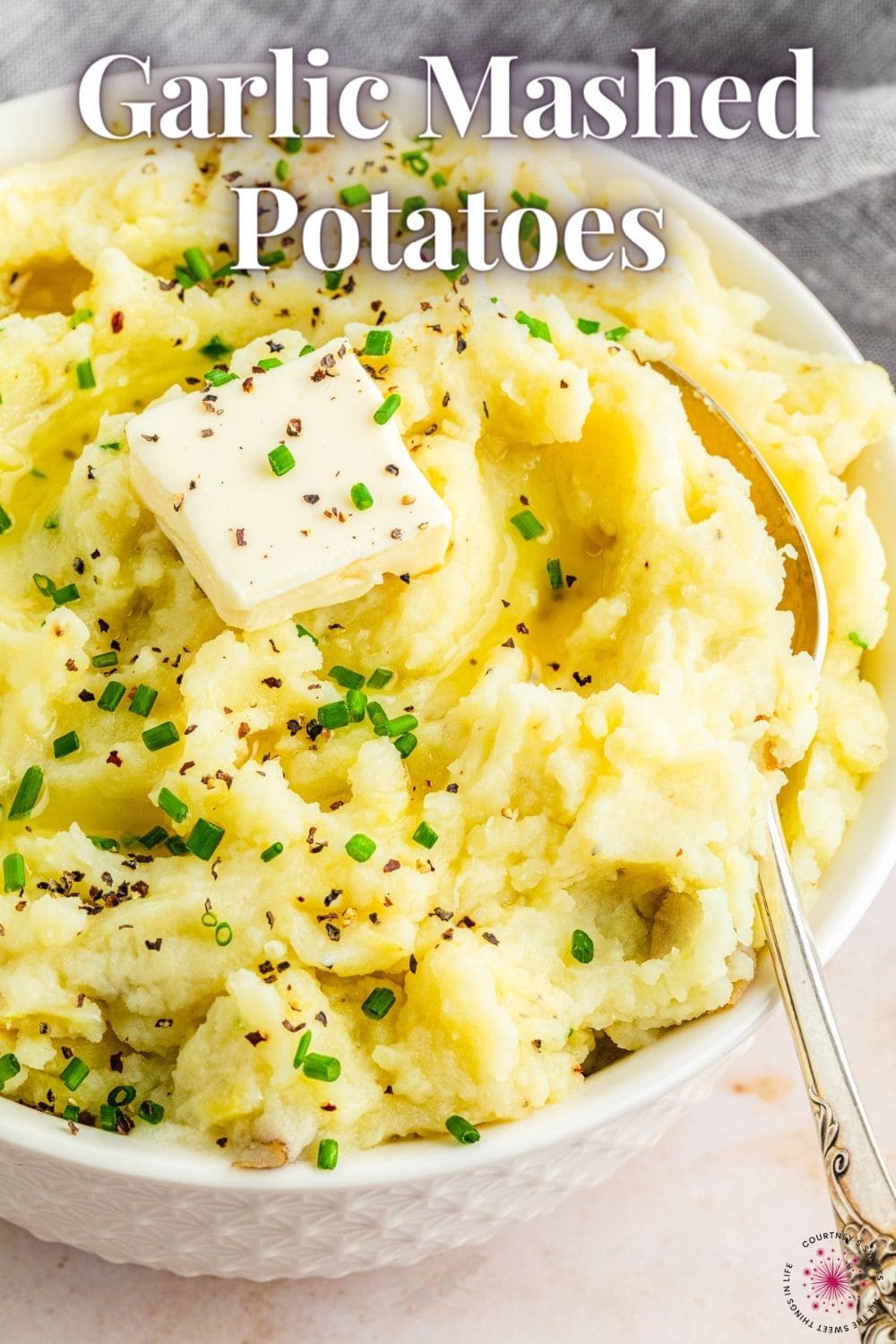 text on image saying garlic mashed potatoes -garlic mashed potatoes in a white bowl with a pat of dairy free butter and chives as garnish and a spoon to the right inside the bowl.