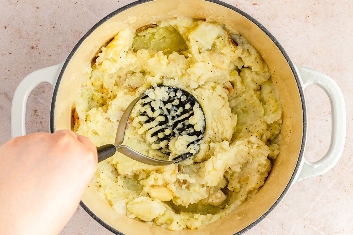 hand holding a potato masher mashing potatoes in a dutch oven.