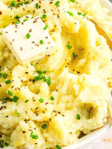 close up of garlic mashed potatoes in a white bowl with a pat of dairy free butter and chives as garnish and a spoon to the right inside the bowl.