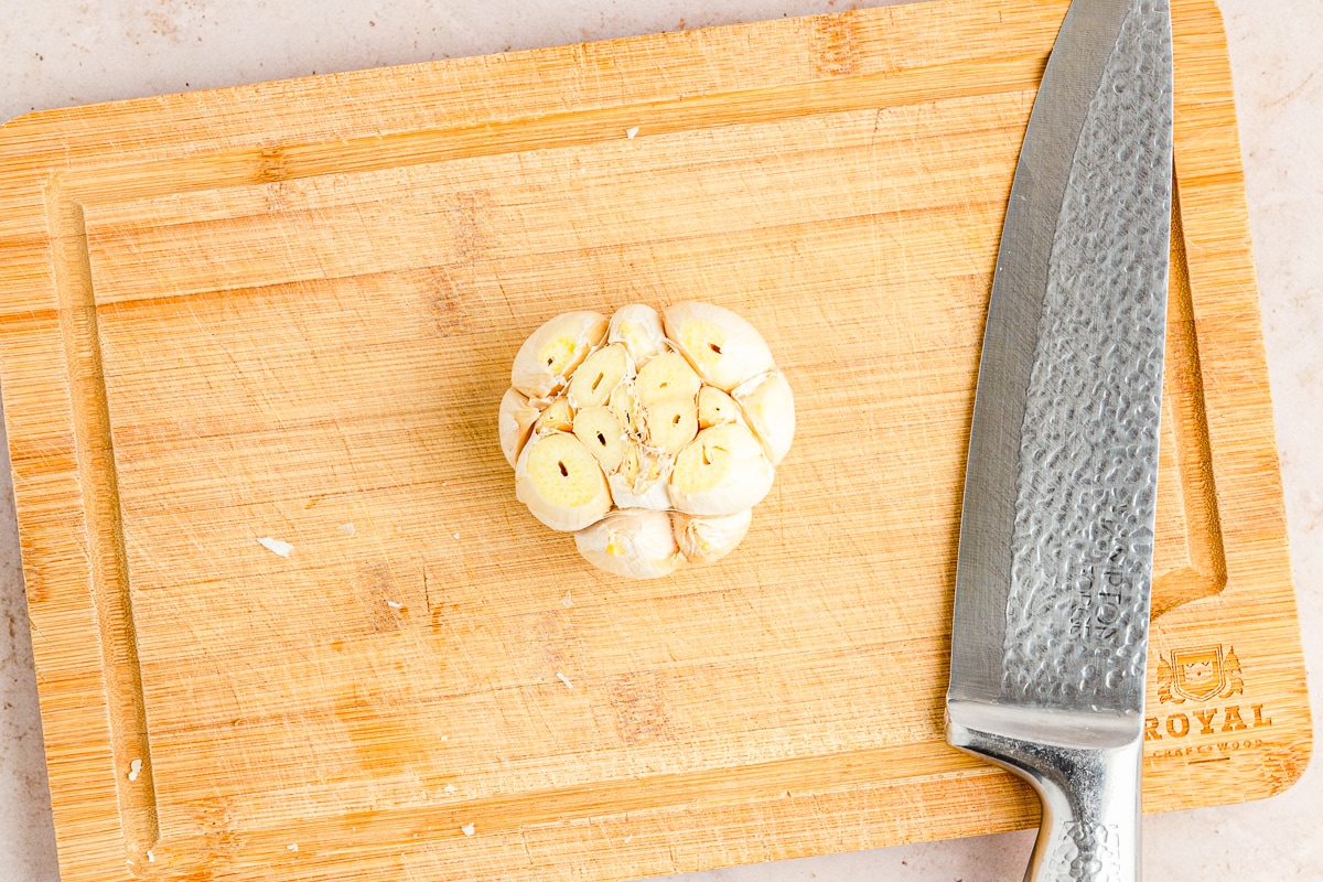 garlic bulb with top sliced off on a cutting board with a chef's knife to the right.