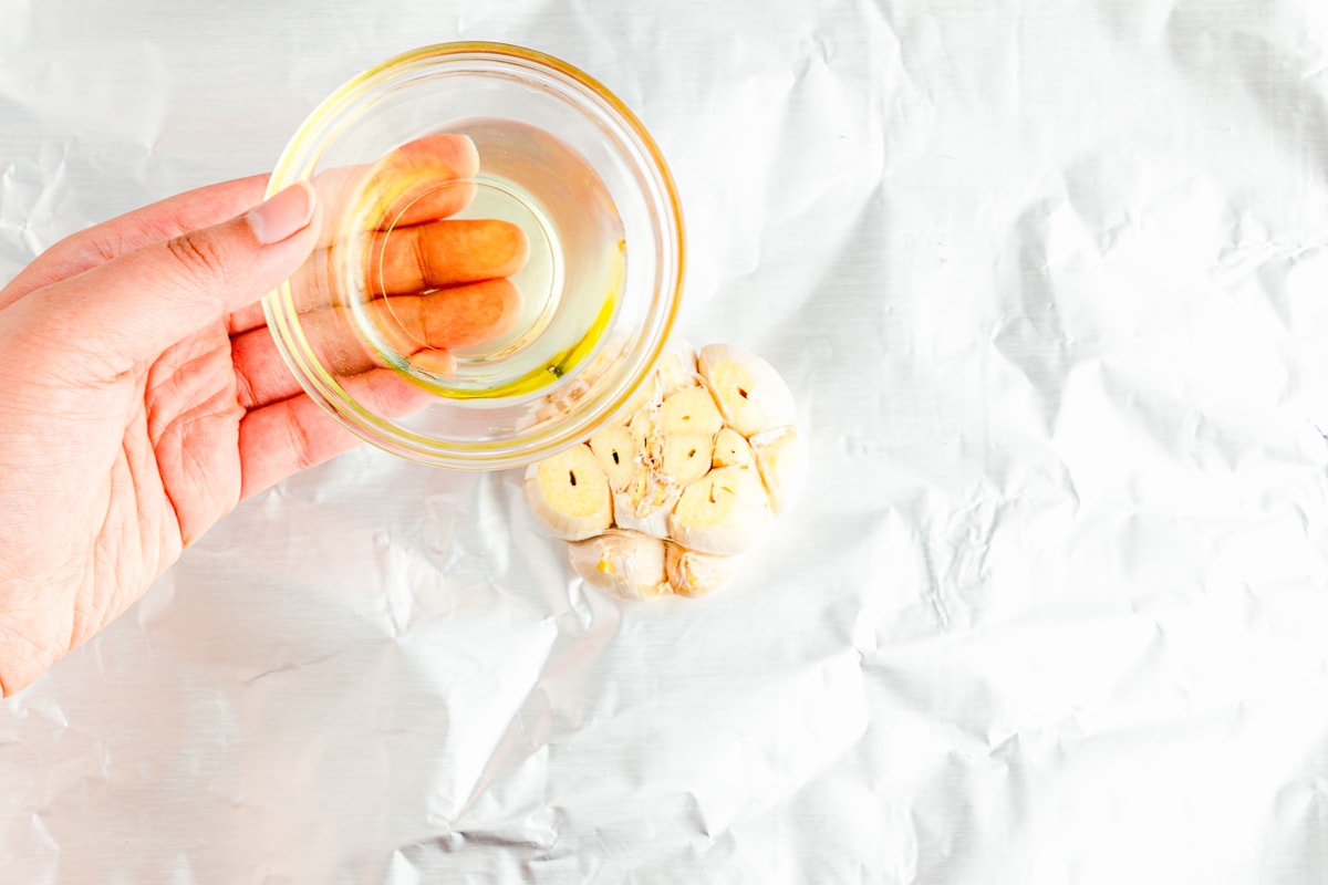 hand holding a small glass bowl of oil over a sliced open garlic bulb on tin foil.