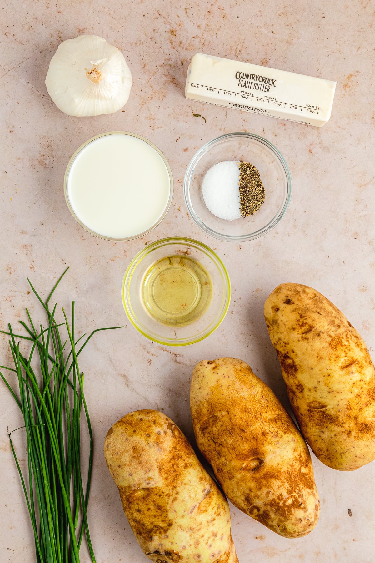 3 russet potatoes, a bunch of chives, small glass bowls filled with oat milk, olive oil, salt and pepper, a whole garlic bulb, a stick of country crock plant butter overhead photo.