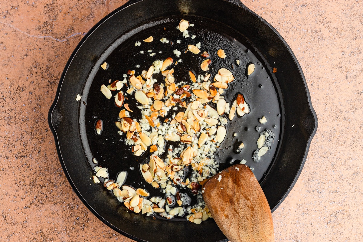 wooden spatula tossing slivered almonds and garlic in a cast iron skillet with oil.