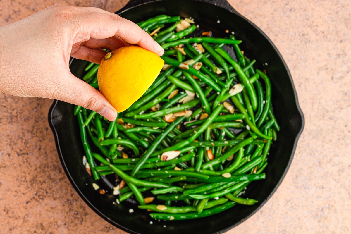 hand adding fresh lemon juice to green beans in a cast iron skillet.