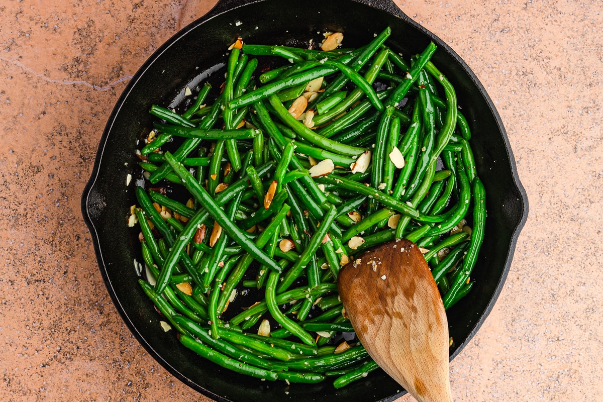 wooden spoon tossing green beans almondine in a cast iron skillet.