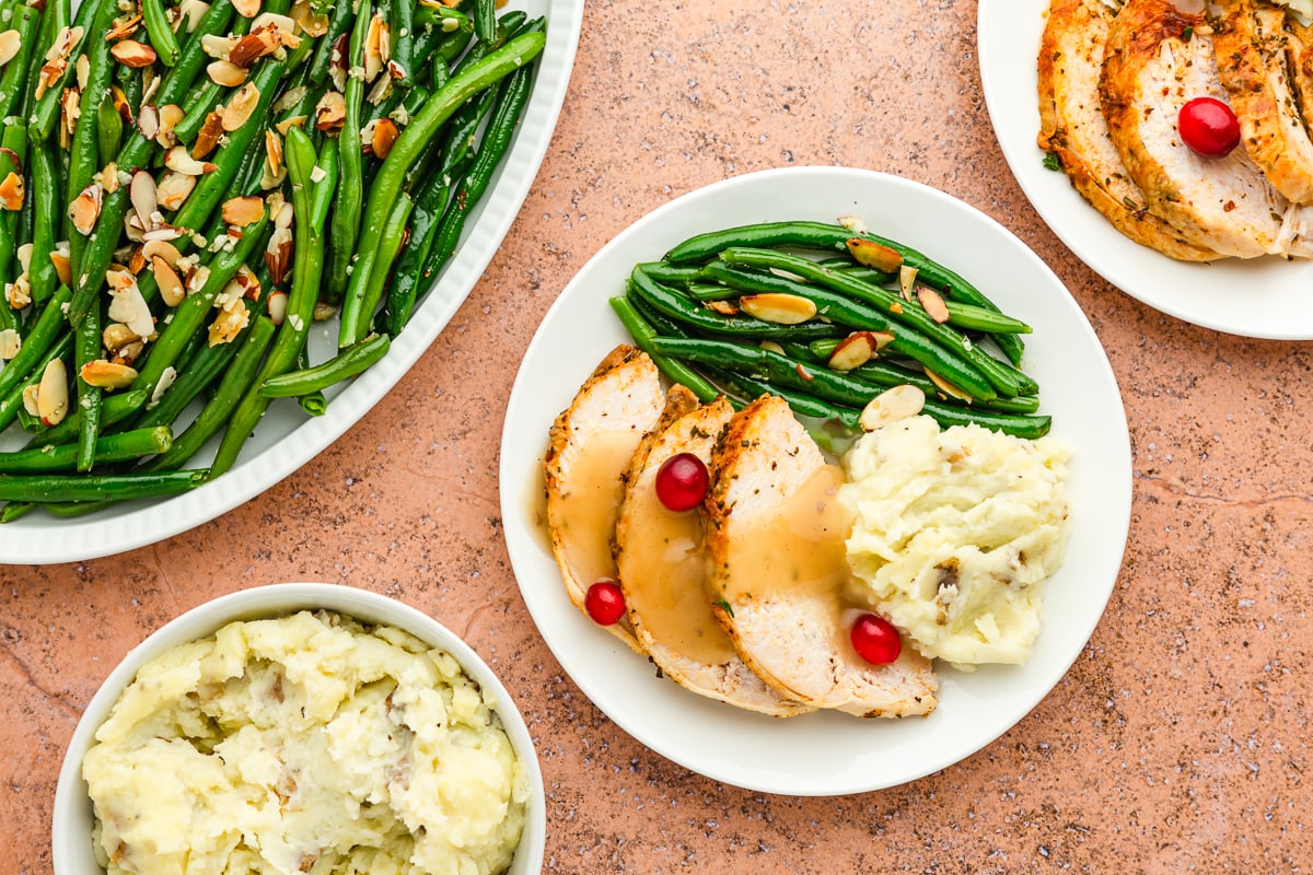 green beans almondine on a white plate with roasted turkey breast slices, cranberries, and mashed potatoes with gravy.