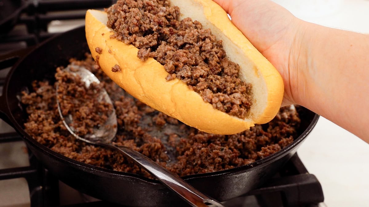 hand holding a ground beef cheesesteak above cast iron skillet.