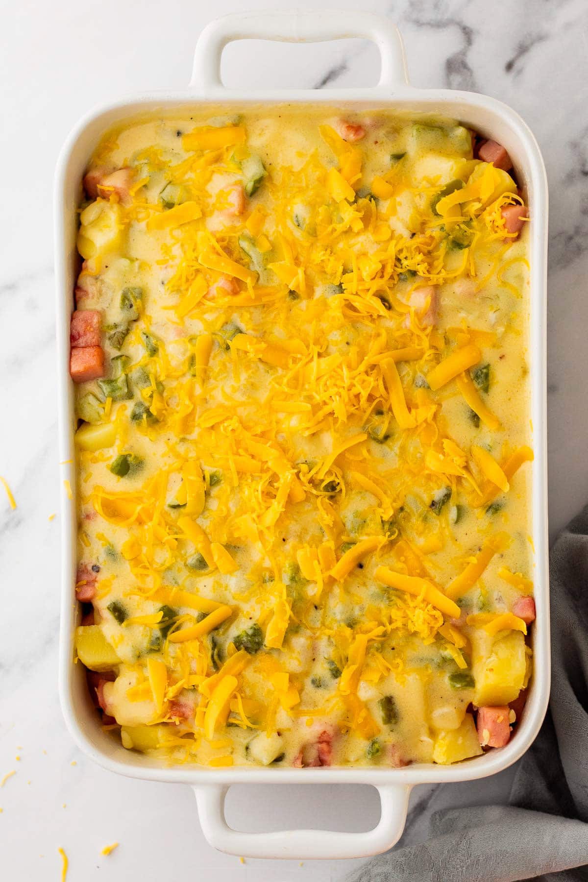 cheese sauce and fresh shredded cheese on top of ham and potatoes in a casserole dish.