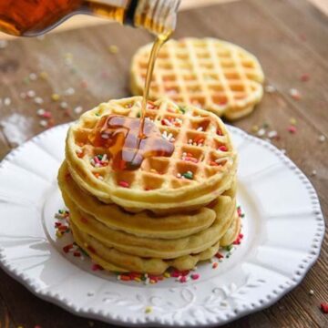 homemade waffles stacked on a plate with maple syrup being drizzled on top.