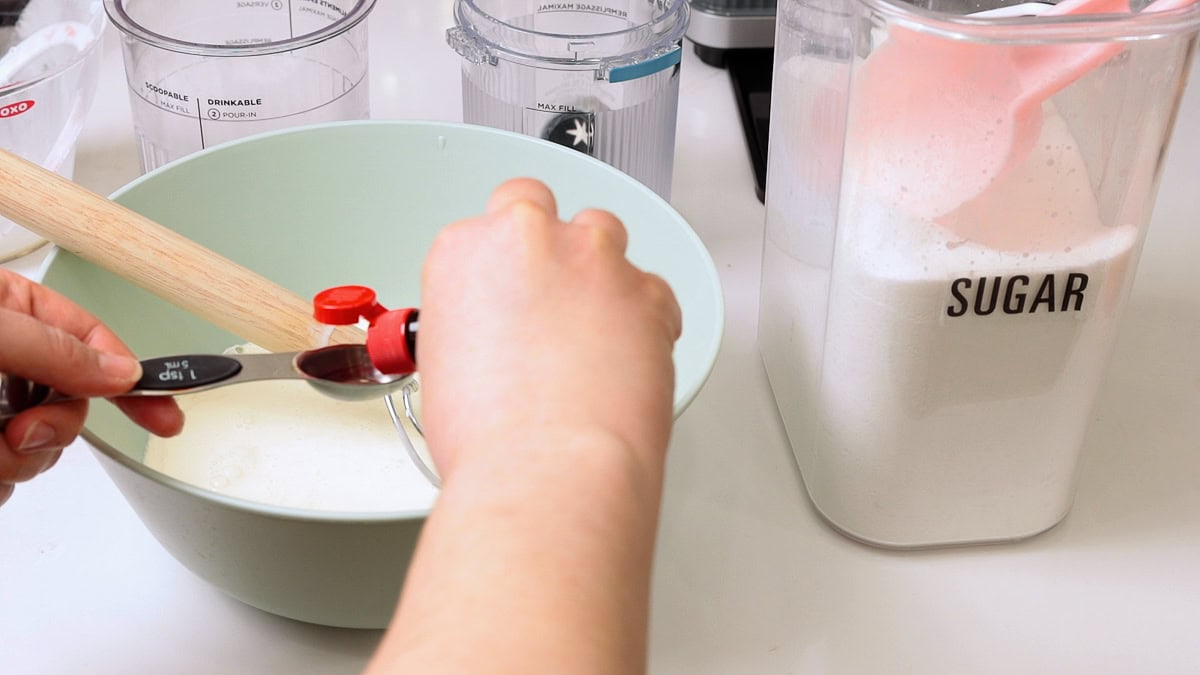 bowl with ingredients for ice cream with hand holding mint extract and pouring it into a measuring spoon.