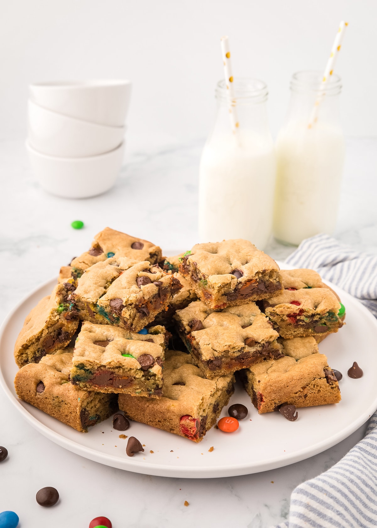 stacked m&m cookie bars on a white plate. with milk in glasses in the background.