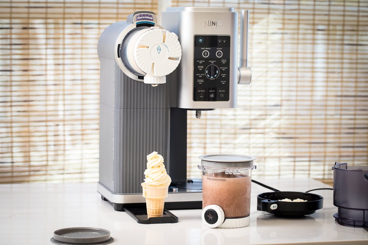 photo of ninja swirl machine on a countertop with an ice cream cone with vanilla soft serve is on the drip tray and a ninja swirl pint is filled with chocolate ice cream recipe ready to blend.