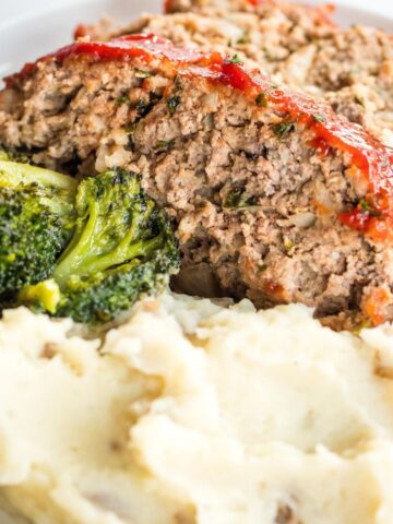 2 slices of meatloaf on a white plate with mashed potatoes and broccoli.