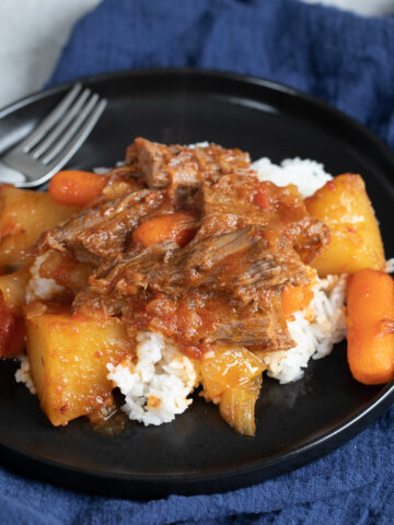 passover brisket with carrots and potatoes over white rice on black plate with navy blue napkin underneath