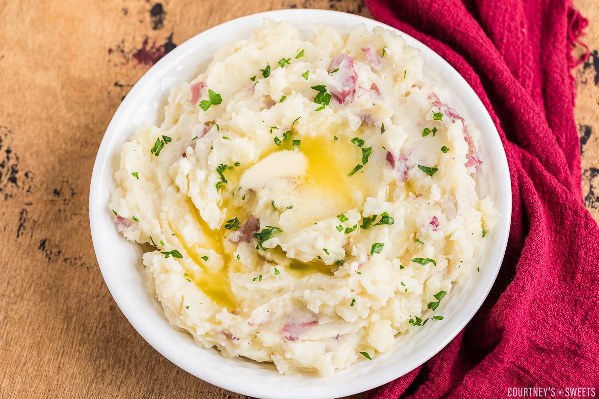 red skin mashed potatoes with melted butter in a white bowl on a dark backdrop with a red napkin to the right side.