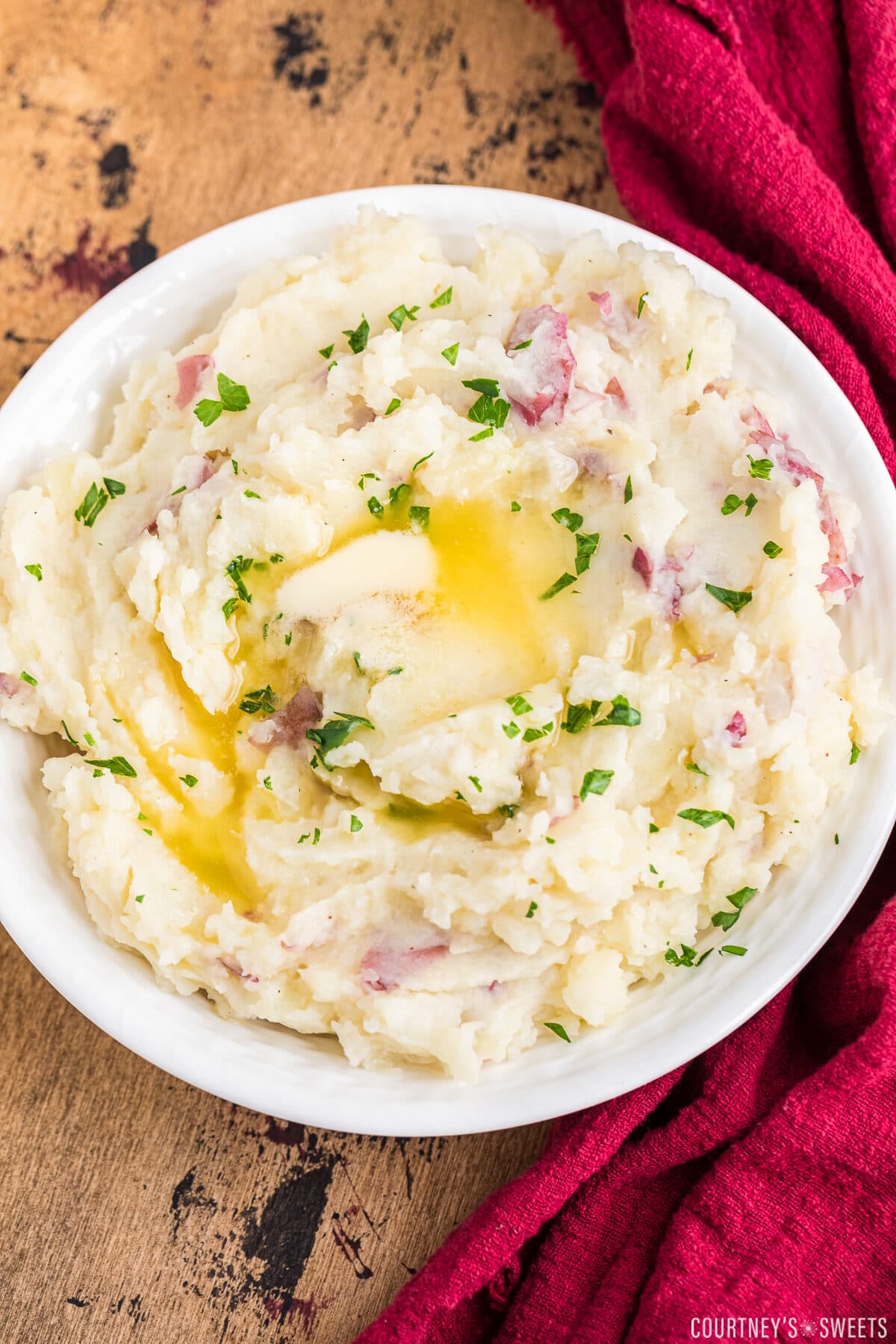 red skin mashed potatoes with melted butter in a white bowl on a dark backdrop with a red napkin to the right.