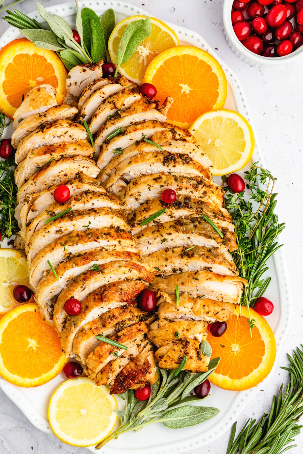 sliced roast turkey breast on a white platter with citrus slices, fresh herbs, and cranberries as garnish.