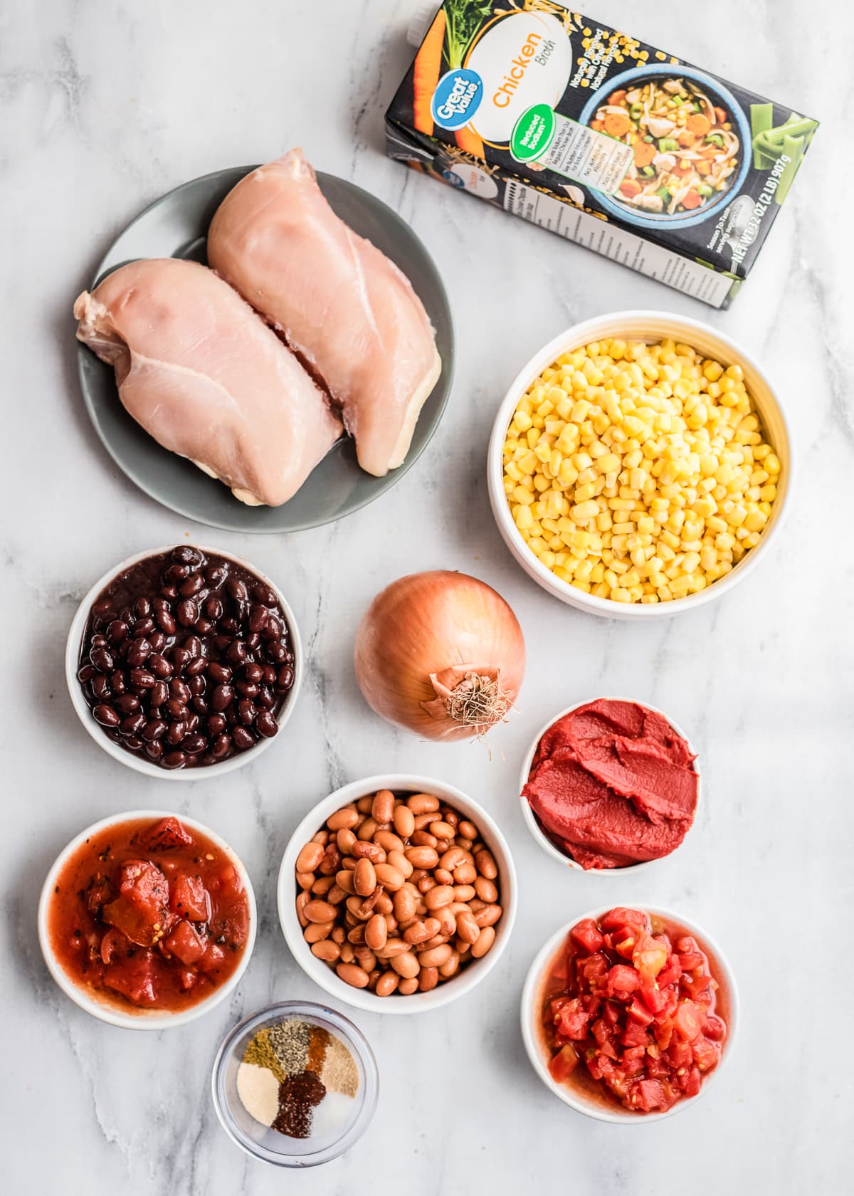 chicken breast on a plate with bowls full of ingredients for chicken taco soup like tomatoes, beans, tomato paste, seasonings, corn, an onion and chicken broth.