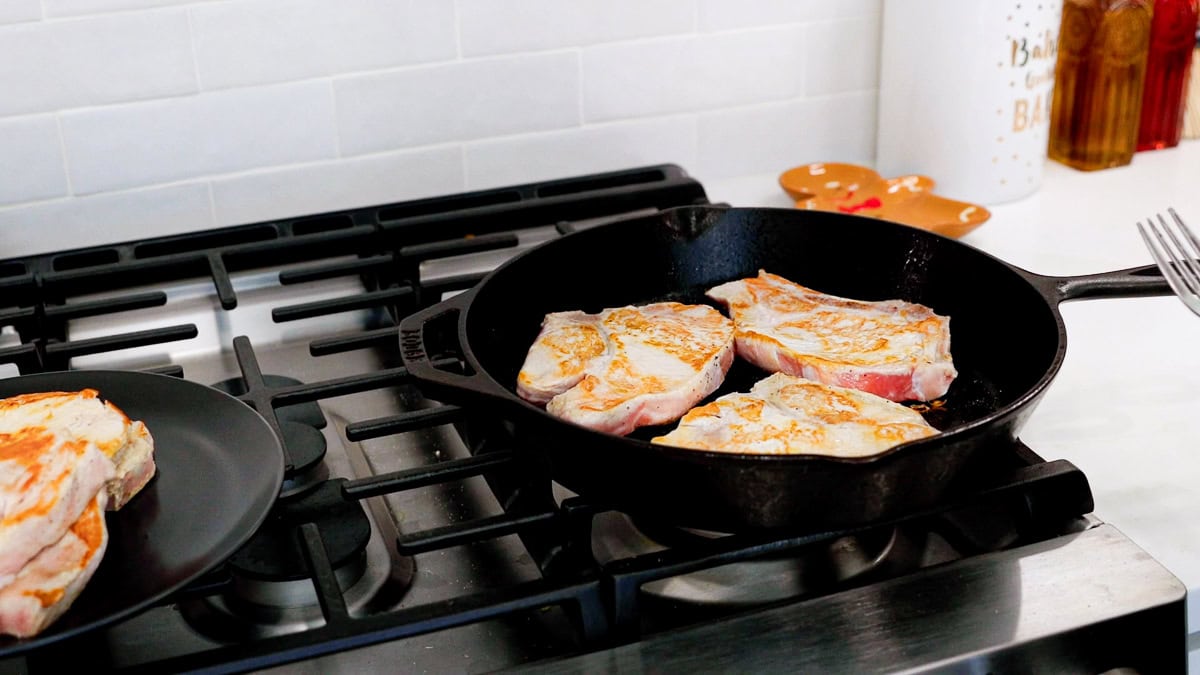 bone in pork chops browning on a cast iron skillet with browned pork chops on a plate to the left.