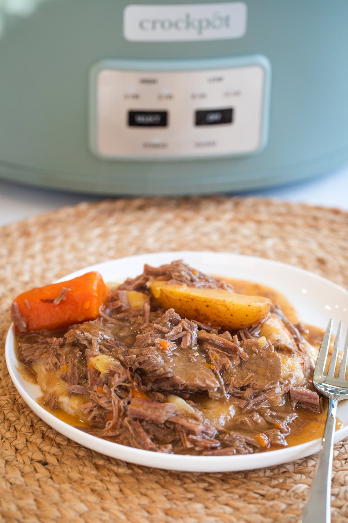 slow cooker top round roast on a plate over mashed potatoes with carrots, potatoes, and gravy in front of a crockpot.