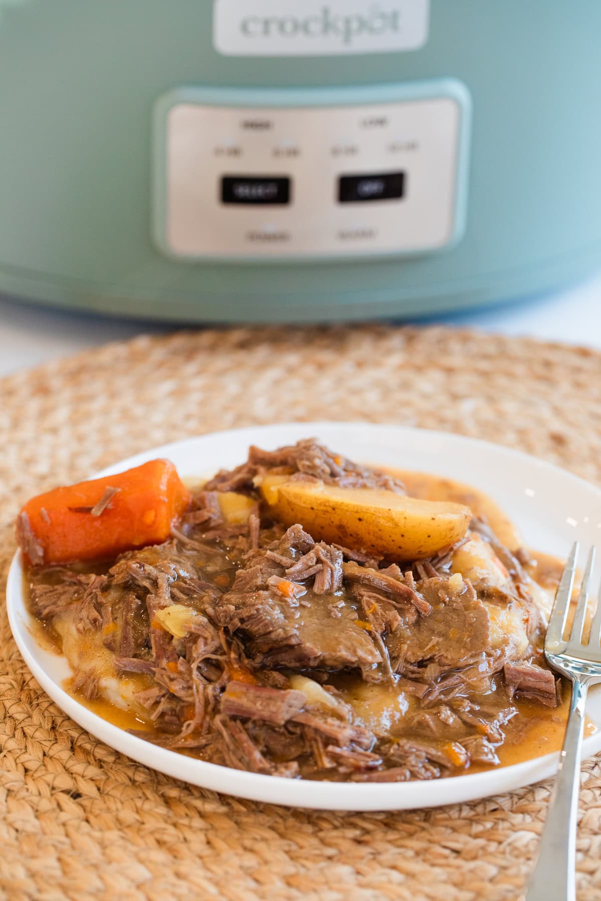 slow cooker top round roast on a plate over mashed potatoes with carrots, potatoes, and gravy in front of a crockpot.