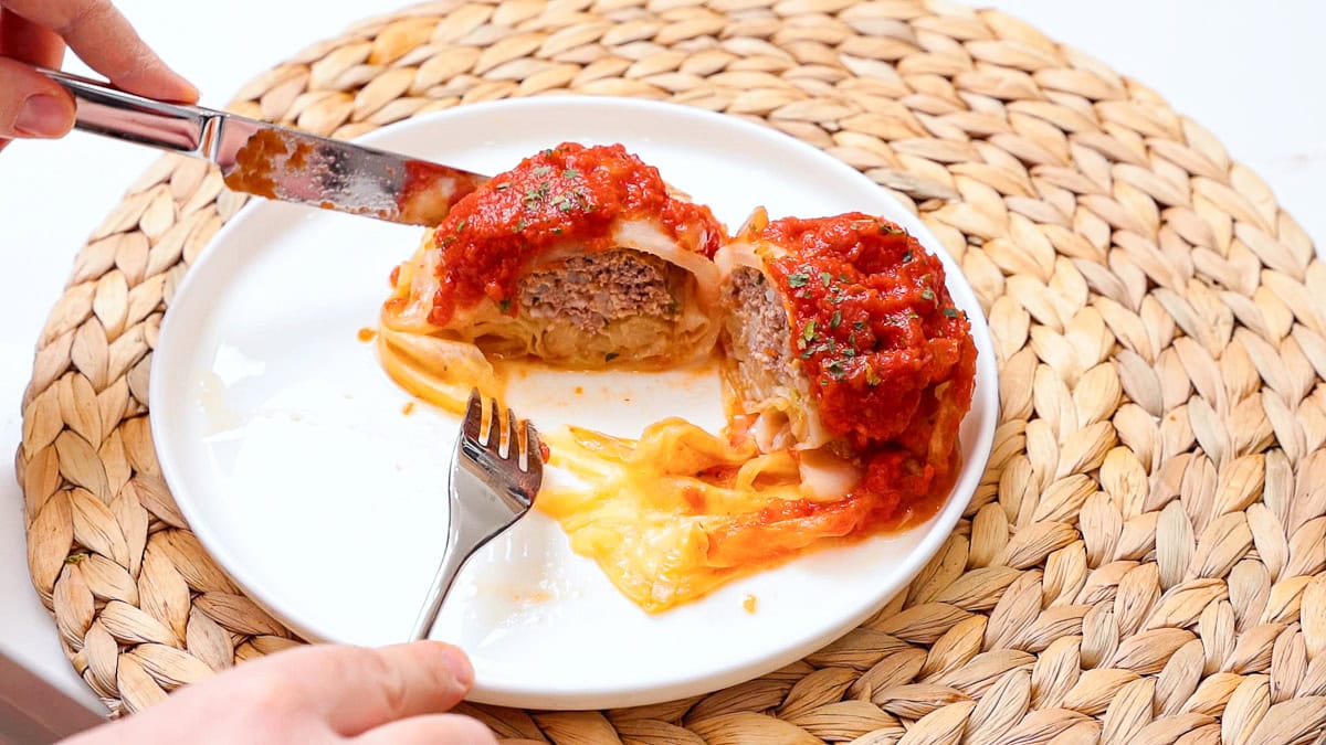 stuffed cabbage rolls on a white plate cut open to show the inside with knife, fork, and hand in photo.