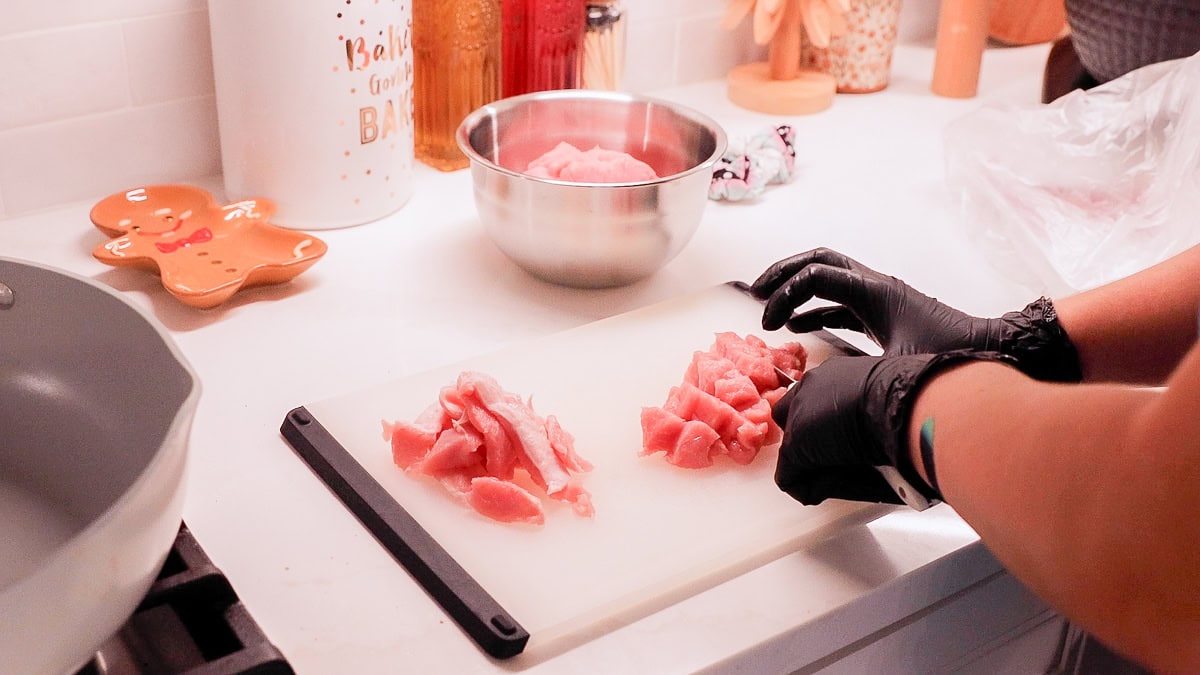 hands in black gloves cutting pork chops into bite size pieces on a cutting board.