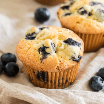 two homemade blueberry muffins on a beige napkin with blueberries around them.