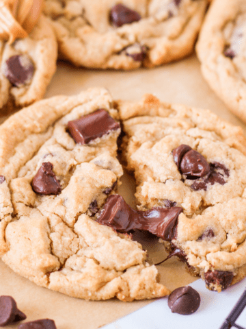 cropped photo of peanut butter chocolate chip cookies on a piece of parchment paper and a spoonful of peanut butter in the background, focus is on cookie torn in half slightly.